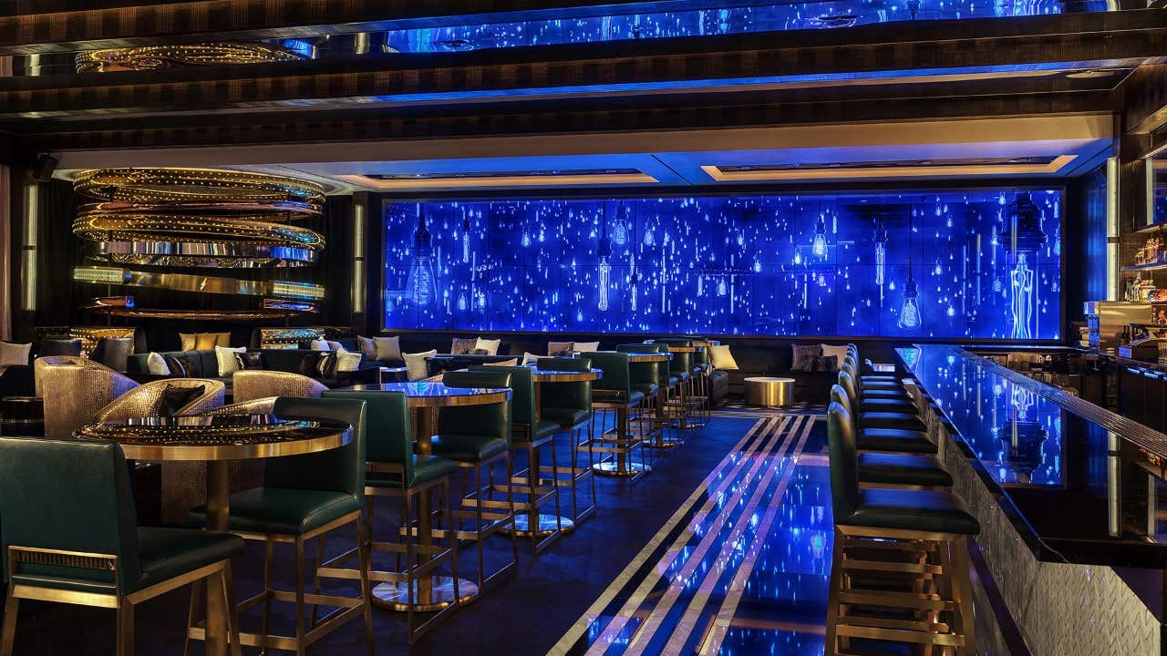 Modern, stylish bar with blue lighting, sleek seating, mirrored ceiling, and a large digital display on the back wall.