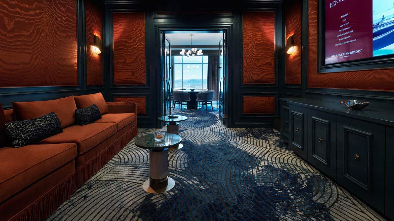 A hotel movie theater room with a long couch on the left, a large wall-mounted TV on the right, and a set of doors in the middle leading to a living room.