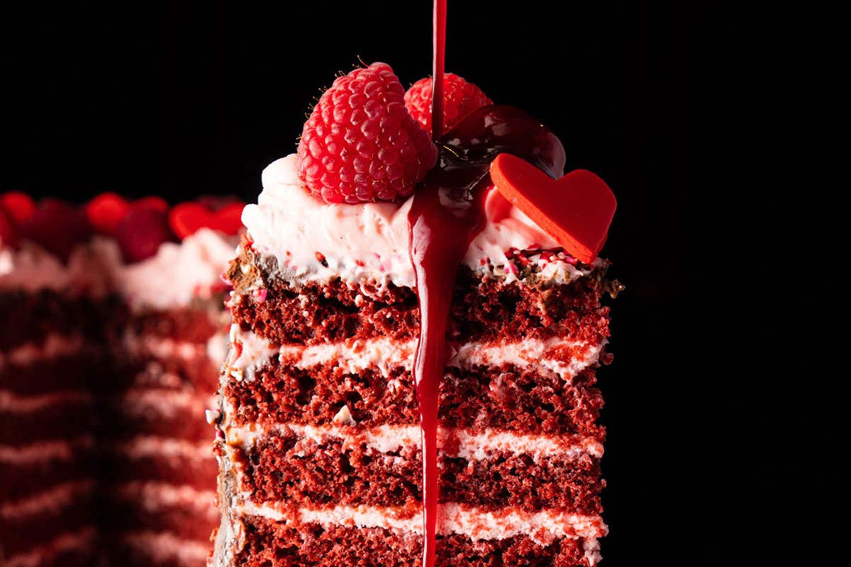 A slice of 20 Layer Red Velvet Cake topped with raspberry, red candied hearts, and red syrup. 
