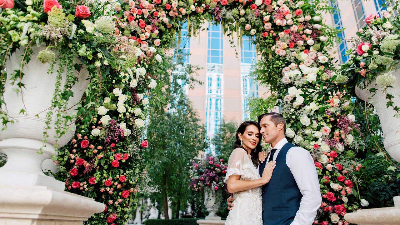 A couple in a wedding dress and suit embrace in front of an outdoor flower arch.