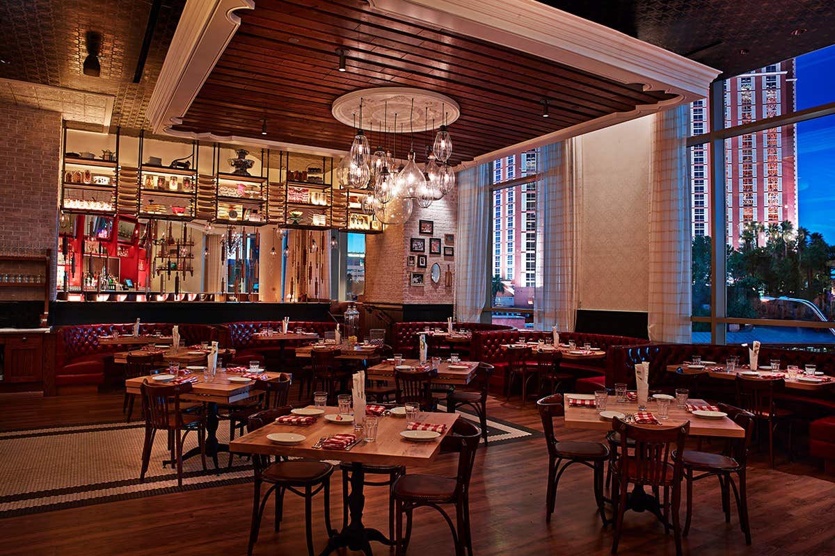 A chic restaurant with wooden tables, chairs, chandeliers, and a well-lit bar, large windows showing a cityscape at dusk.