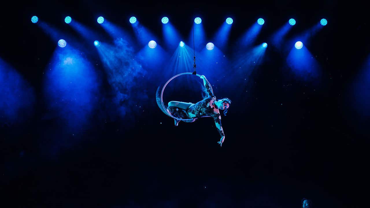 Shin Lim looking up to a woman dressed in black lace hanging onto a hoop that is suspended in the air.