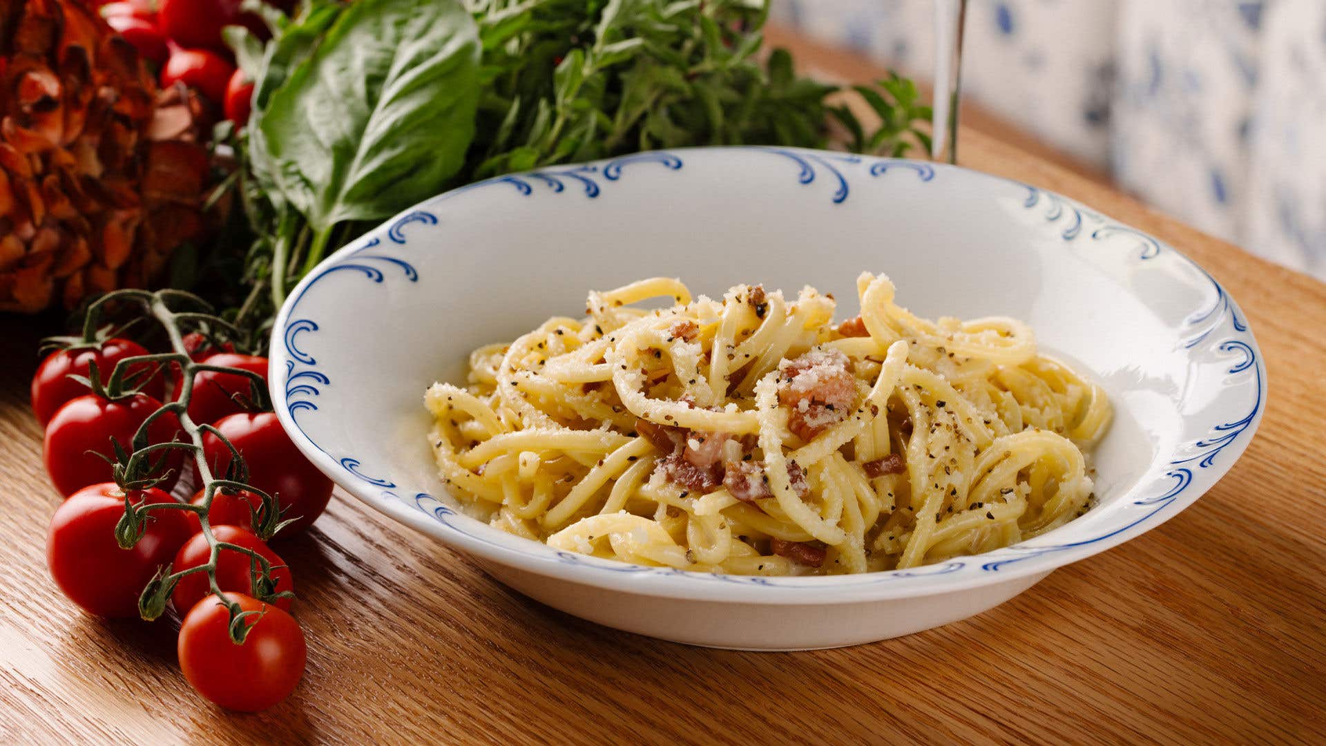 Pasta dish in a white bowl on a wood table with tomatoes and assorted vegetables nearby. 