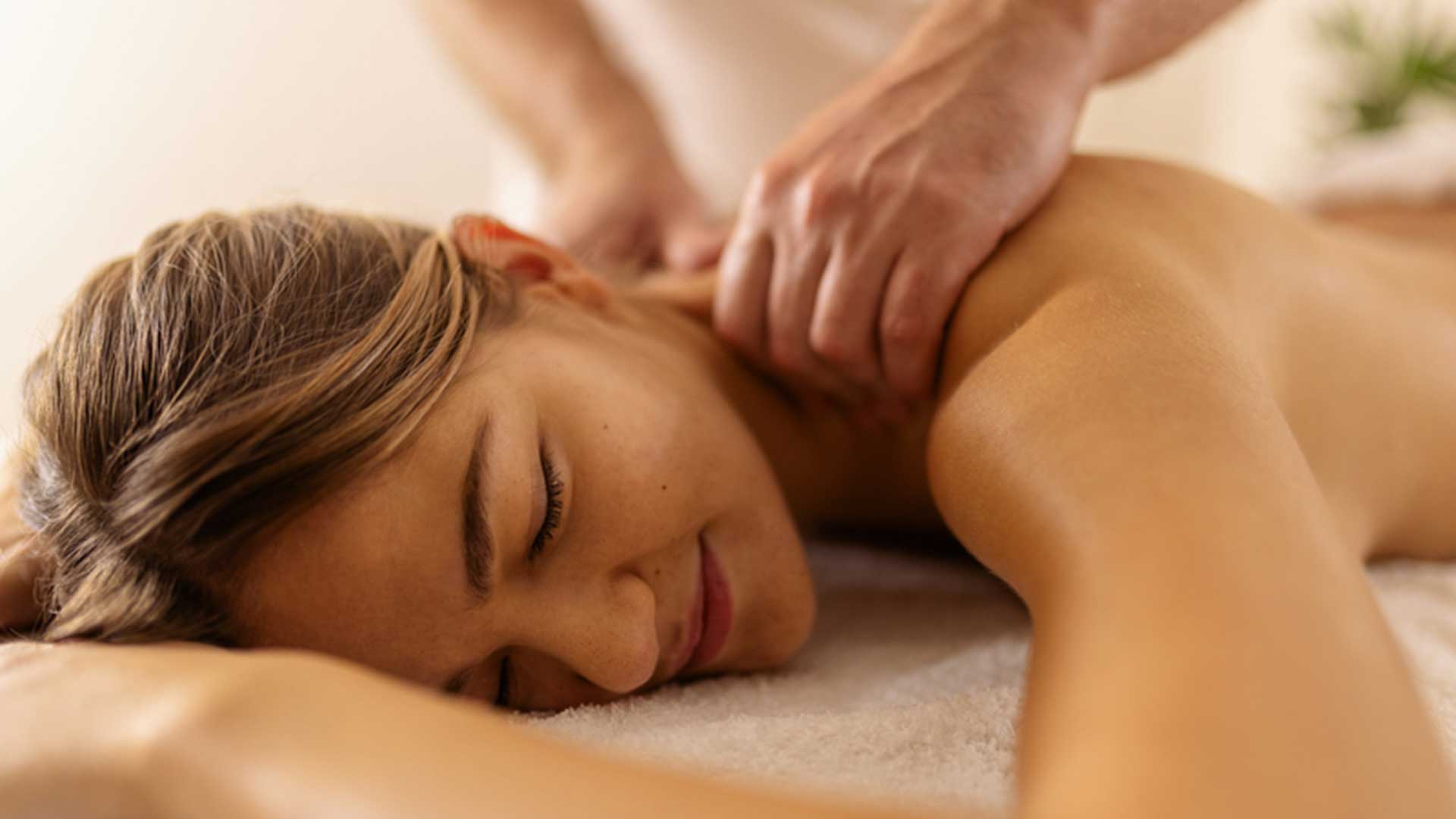 A woman lays face-down on a massage table while a masseuse massages her shoulders.