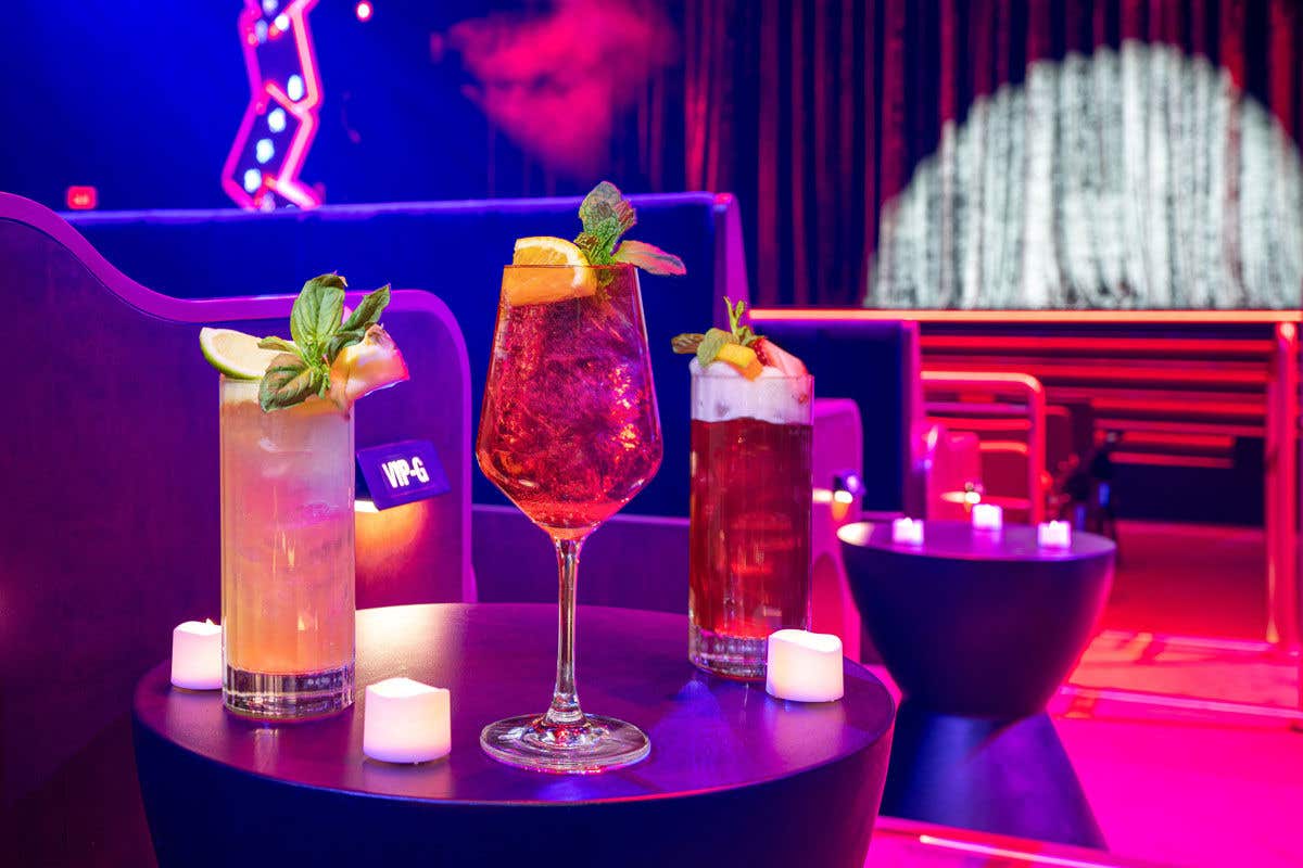 Three cocktails on a table in a dimly lit lounge with colorful lighting and booth seating.