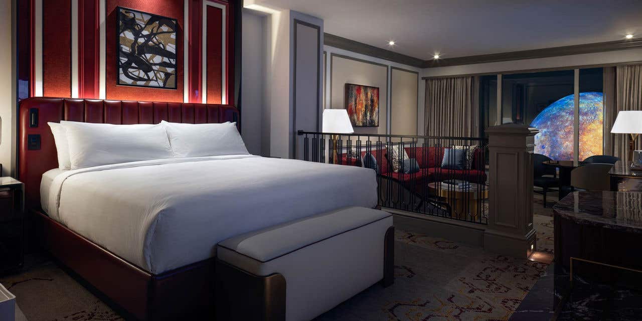 Luxury King Suite bedroom with a bench seat at the foot of the bed. In the background is a sunken living room with an armchair, couch, coffee table, dining table with chairs, lamps, and a nighttime view of Sphere at The Venetian outside the window.