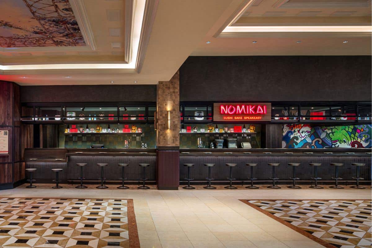 Modern bar with a long counter, black stools, shelves of bottles, and a neon sign reading “NOMIKAI Sushi Sake Speakeasy” above the bar.