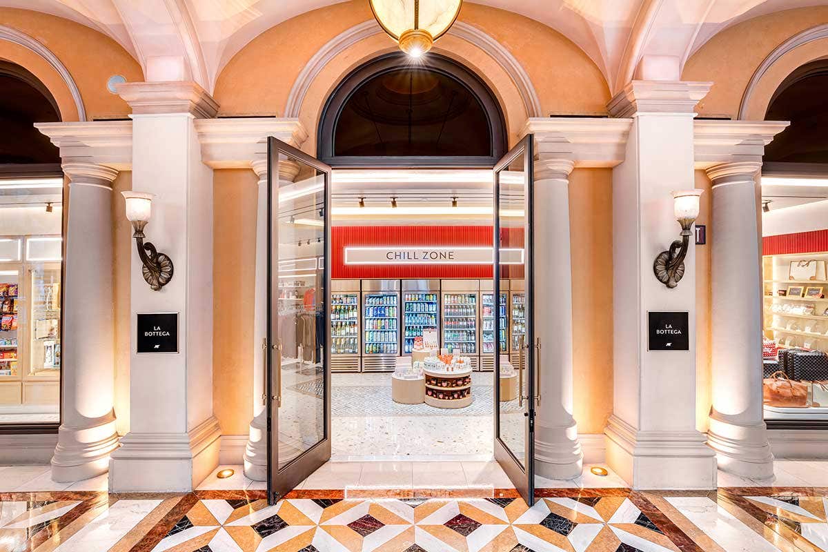 An open doorway to a store containing varieties of items, from a fridge in the back containing drinks, to clothing and baggage to the right